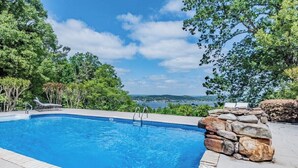 A swimming pool with a stone wall and lounge chairs, surrounded by trees and a view of the landscape. - Spacious 5BR estate with Jacuzzi, pool, patio, and full kitchen. (Guntersville)