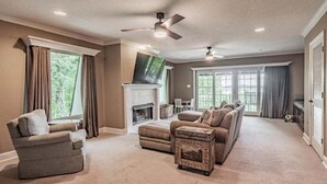 A living room with a fireplace, ceiling fans, and a large window. - Spacious 5BR estate with Jacuzzi, pool, patio, and full kitchen. (Guntersville)
