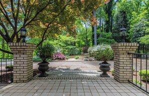 A paved pathway leading to a brick entrance with two lantern-style lights, surrounded by lush greenery and trees. - Spacious 5BR estate with Jacuzzi, pool, patio, and full kitchen. (Guntersville)