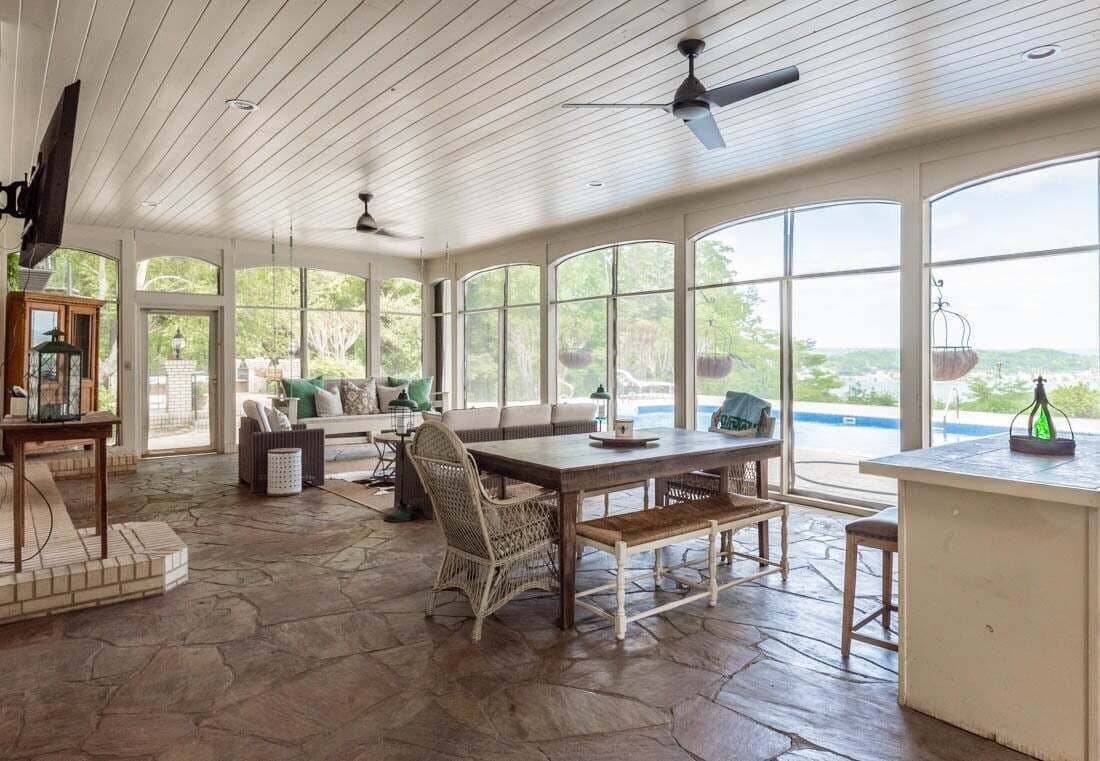 A spacious sunroom with a stone floor, large windows, a dining table, wicker chairs, and ceiling fans.