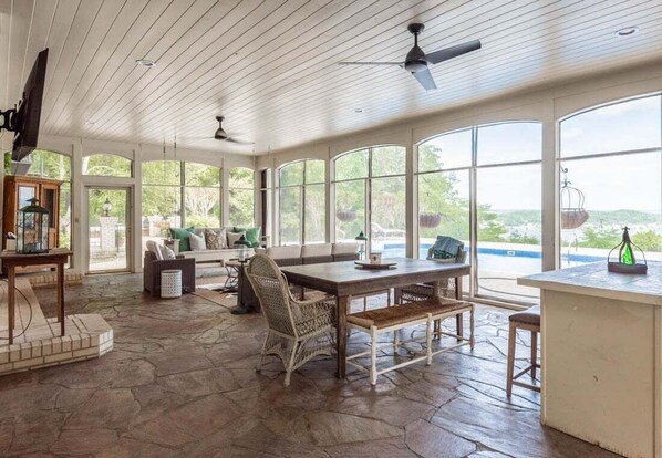 A spacious sunroom with a stone floor, large windows, a dining table, wicker chairs, and ceiling fans.