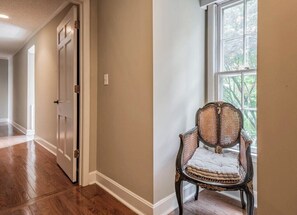 A hallway with wooden flooring, a door, and a chair near a window.
