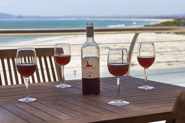 A wooden table with a bottle of red wine and four wine glasses on a beach overlooking the ocean.