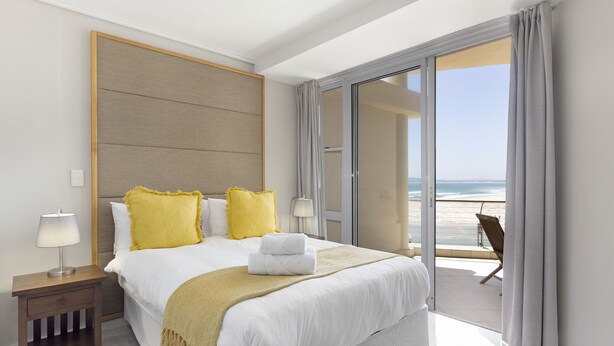 Une chambre d’hôtel avec un grand lit, une table de chevet, une lampe et une vue sur la plage, accessible par une porte-fenêtre coulissante.