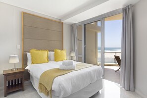 A hotel room with a large bed, a nightstand, a lamp, and a view of the beach through a sliding glass door.