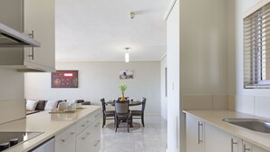 Une cuisine moderne avec des meubles blancs, un coin repas comprenant une table et des chaises, et une grande fenêtre avec des stores.