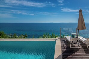 A poolside area with a view of the ocean, a lounge chair, and an umbrella. - Pelagos Blue Zante (Zakynthos)