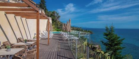 A wooden deck with lounge chairs and tables, overlooking a clear blue sea and a mountainous backdrop.