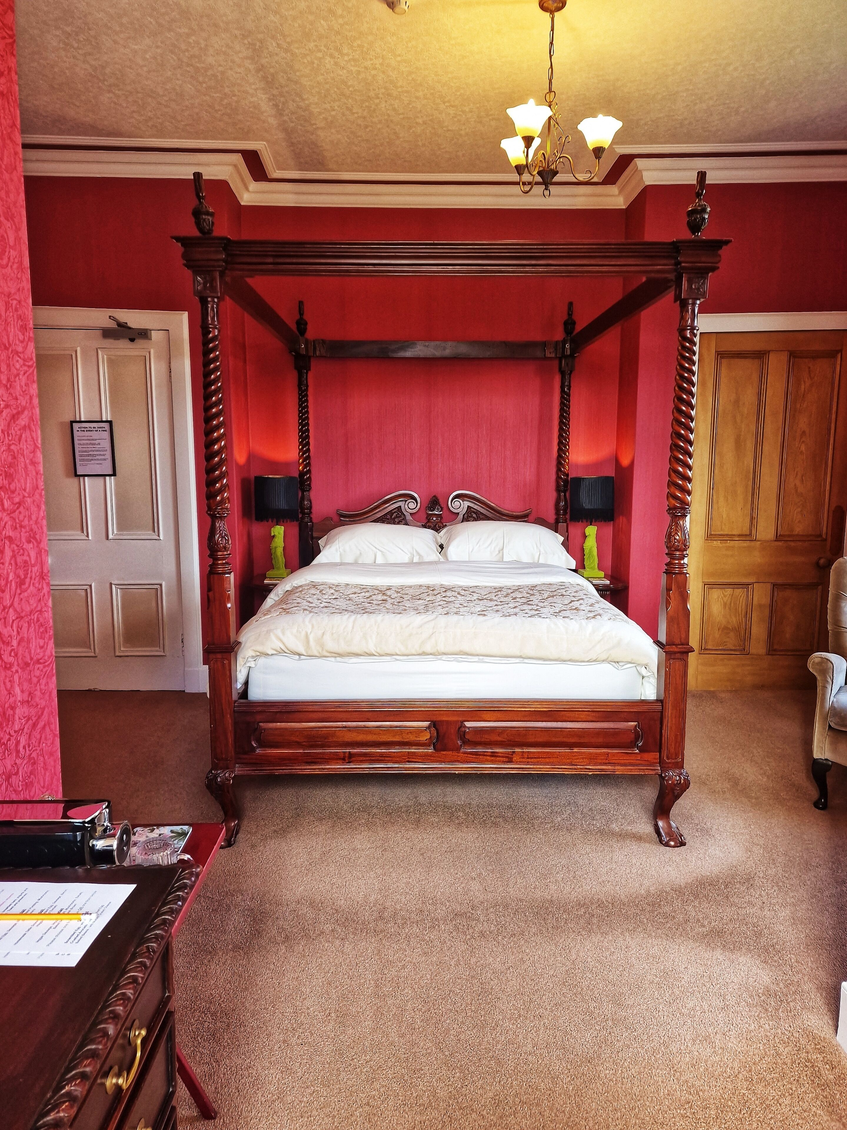 A four-poster bed with a wooden frame and white linens, a chandelier, and a red wall.