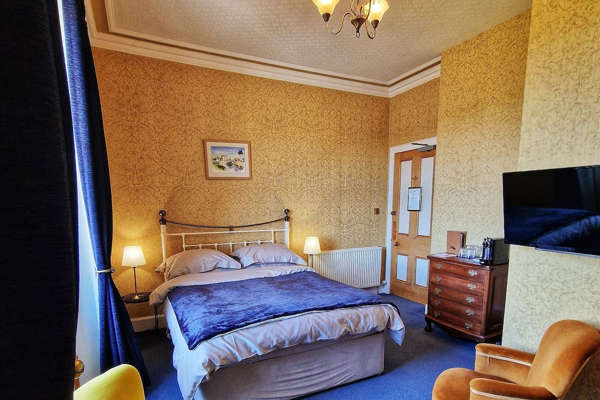 A bedroom with a bed, a chair, a dresser, a television, and a chandelier.