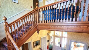 A grand staircase with a wooden handrail and balusters, leading to a lower level with a patterned carpet and a seating area. - The Pines Guesthouse (Elgin)