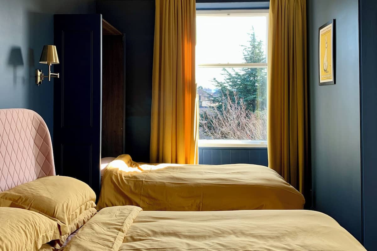 A bedroom with two beds, a window with curtains, a wall-mounted lamp, and a framed picture.