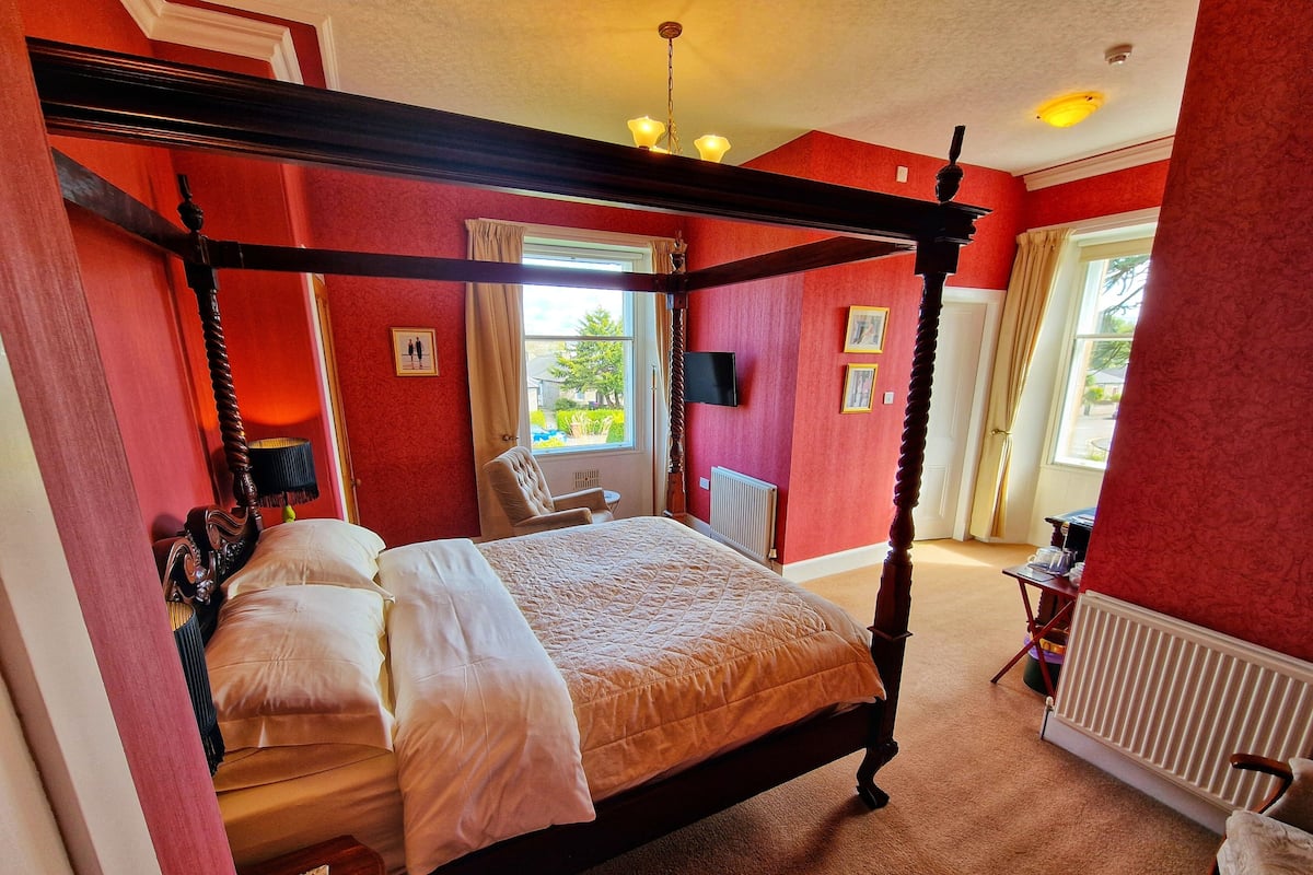 A four-poster bed with a quilt, a chair, a radiator, and a window with curtains.