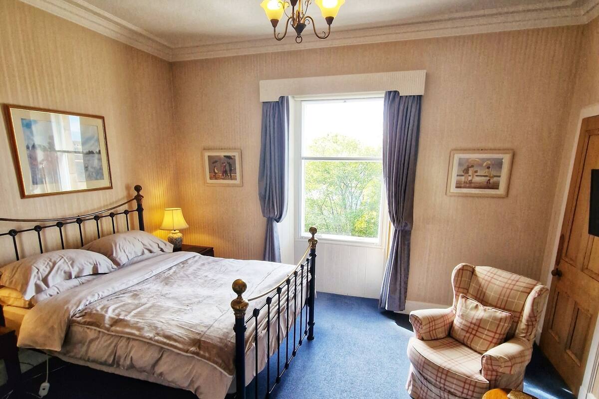 A bedroom with a bed, a chair, a nightstand, a lamp, a window with curtains, and a chandelier.