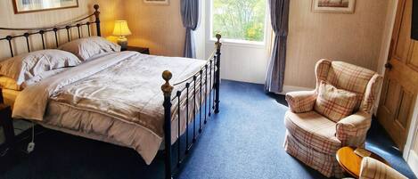 A bedroom with a bed, a chair, a nightstand, a lamp, a window with curtains, and a chandelier.