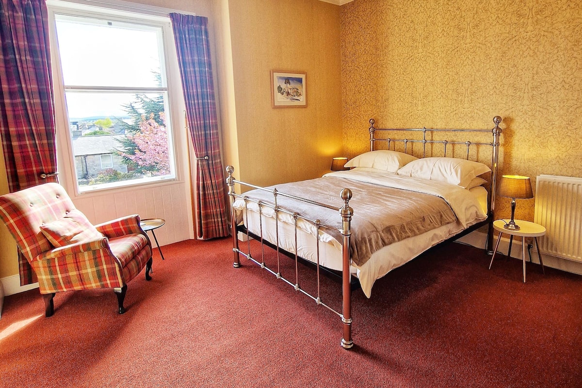 A hotel room with a bed, a chair, a small table, a window with curtains, and a chandelier.