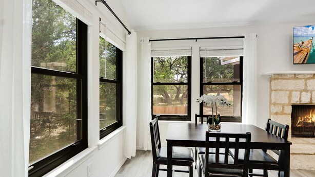 A dining area with a table and chairs, large windows, a fireplace, and a painting on the wall. - Woodacre Cottage I   Minutes to Jacob's Well (Wimberley)