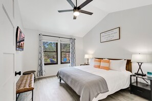 Un dormitorio con una cama, dos mesitas de noche con lámparas, un banco, un ventilador de techo y una ventana con cortinas.