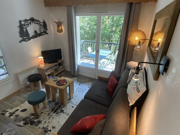 A cozy living room with a sofa, a TV, a small table, and a window overlooking a balcony.