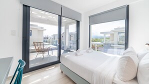 Une chambre moderne avec un grand lit, un balcon avec un fauteuil et une vue sur les immeubles.