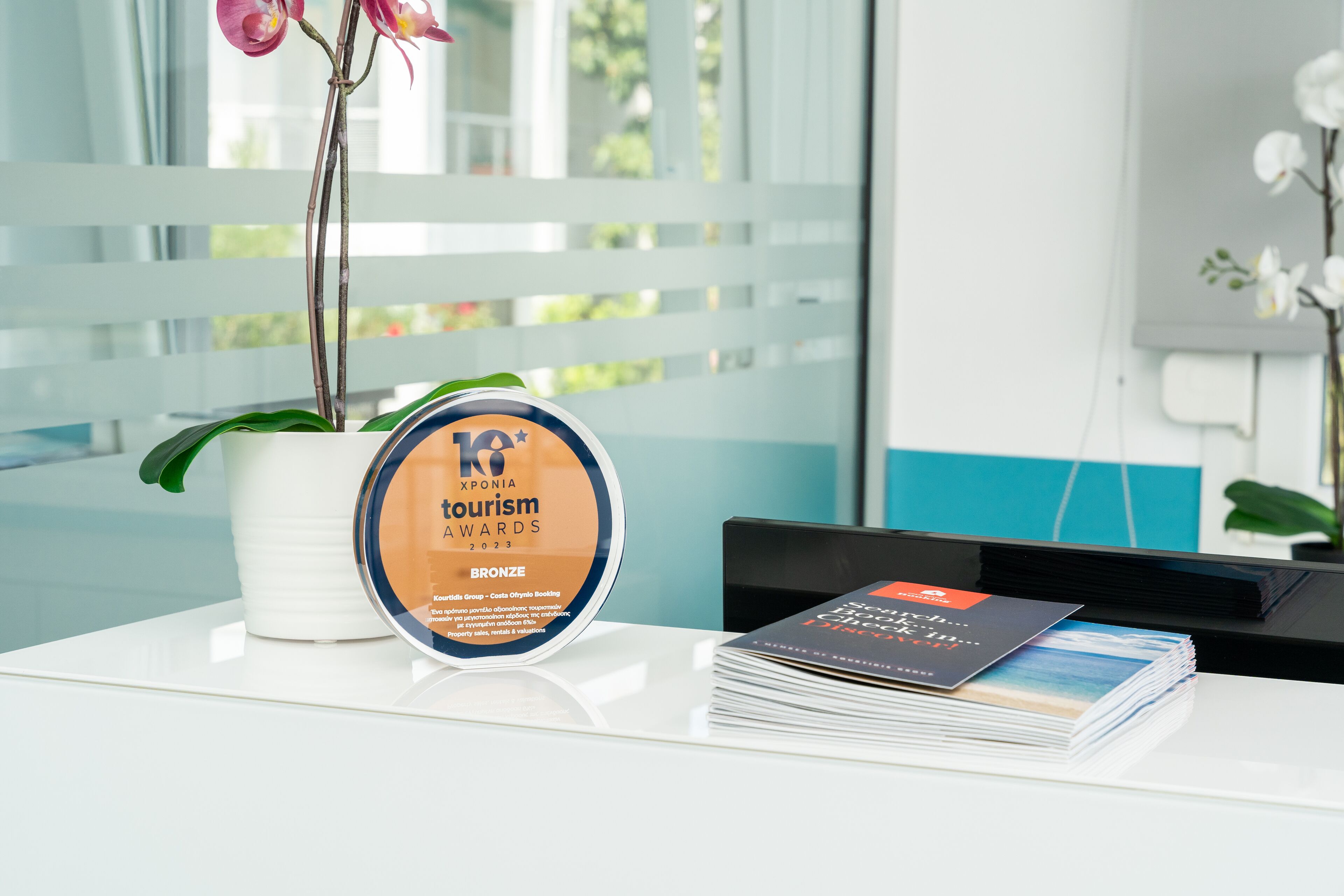 A desk with a trophy for the Xplora Tourism Awards 2023, a stack of brochures, and a potted orchid.