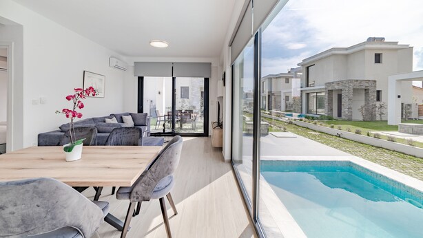 Un salon moderne avec une table à manger en bois, un canapé gris et une vue sur une piscine et les maisons voisines.