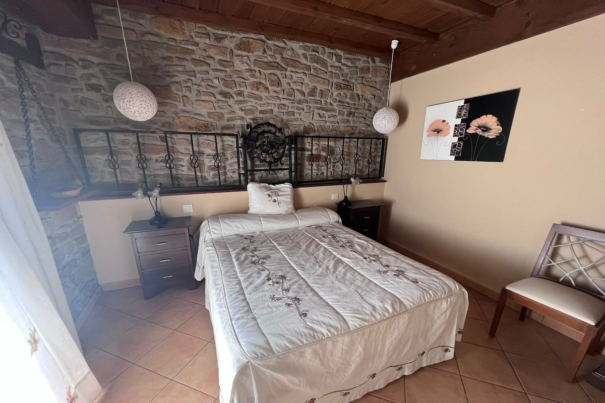 A bedroom with a stone wall, a bed with a floral bedspread, a nightstand, a chair, and a wall-mounted artwork.