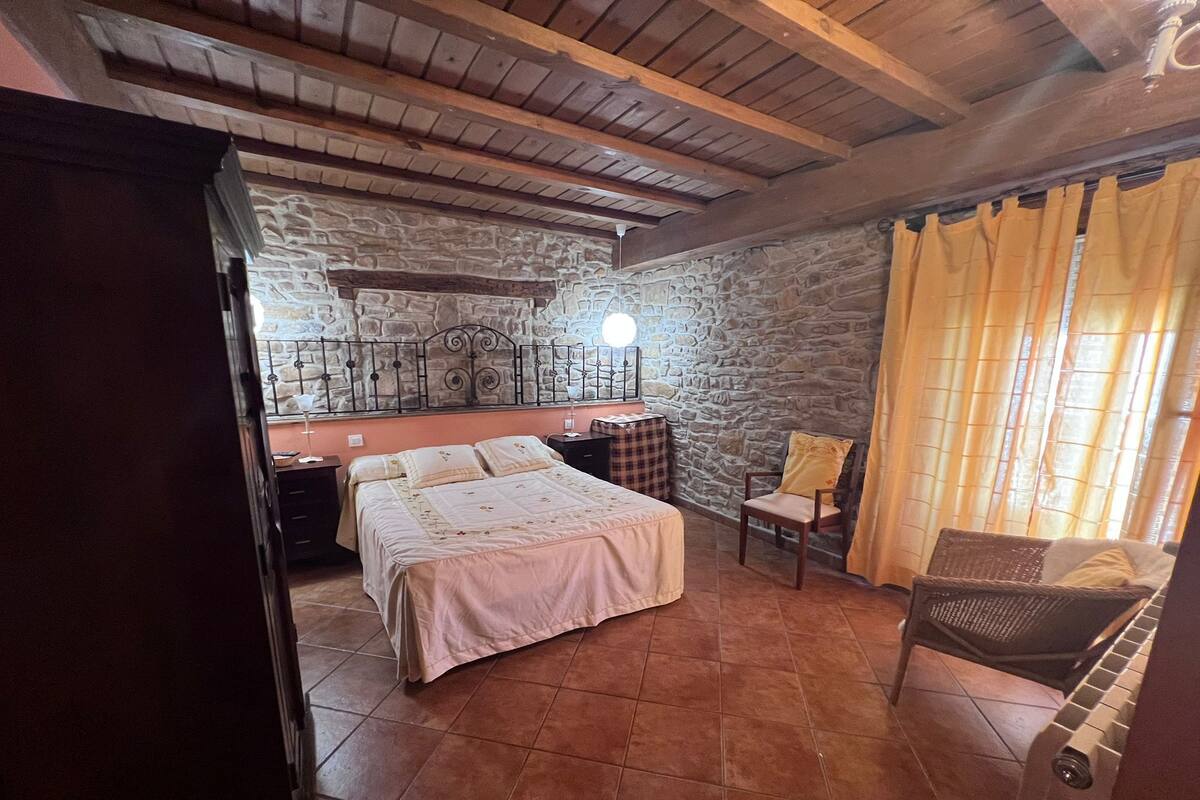 A bedroom with a stone wall, a bed, a chair, and a small table with a lamp.