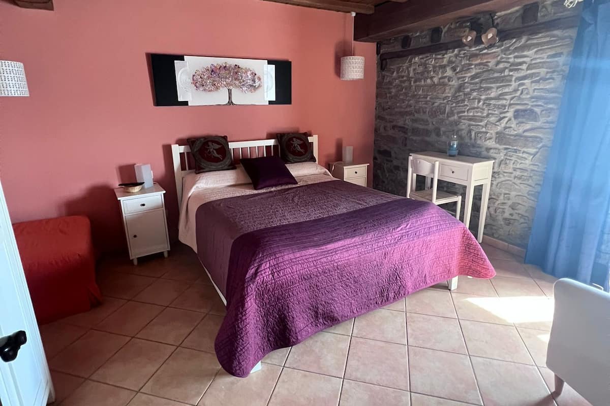 A bedroom with a wooden ceiling, a bed with a purple bedspread, a nightstand, a red chair, and a stone wall.