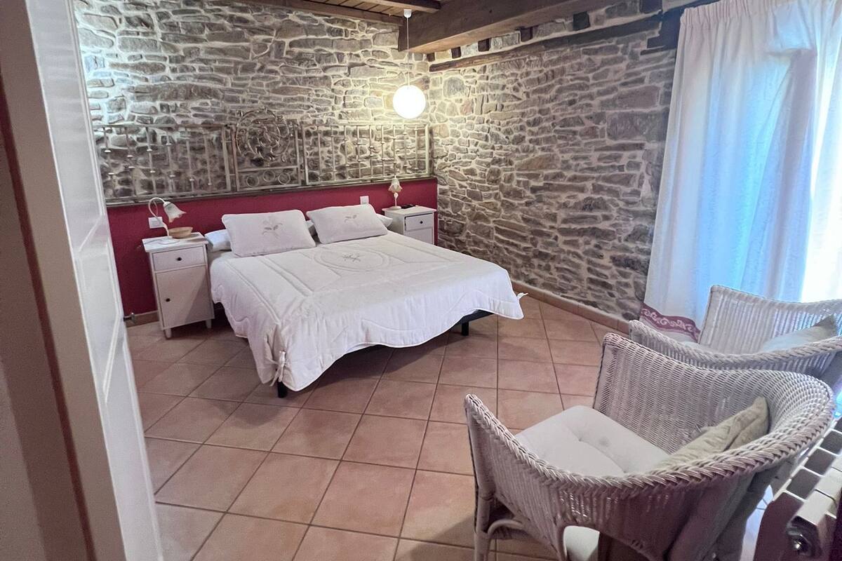 A bedroom with a stone wall, a bed with white linens, a wicker chair, and a hanging light fixture.
