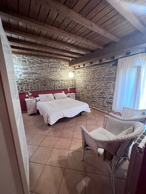A bedroom with a stone wall, a bed with white linens, a wicker chair, and a hanging light fixture. - Hotel rural casa la Juana (Almarza)
