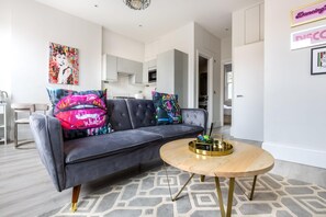 Un salon moderne avec un canapé gris, des coussins colorés, une table basse ronde en bois, et une cuisine en arrière-plan.