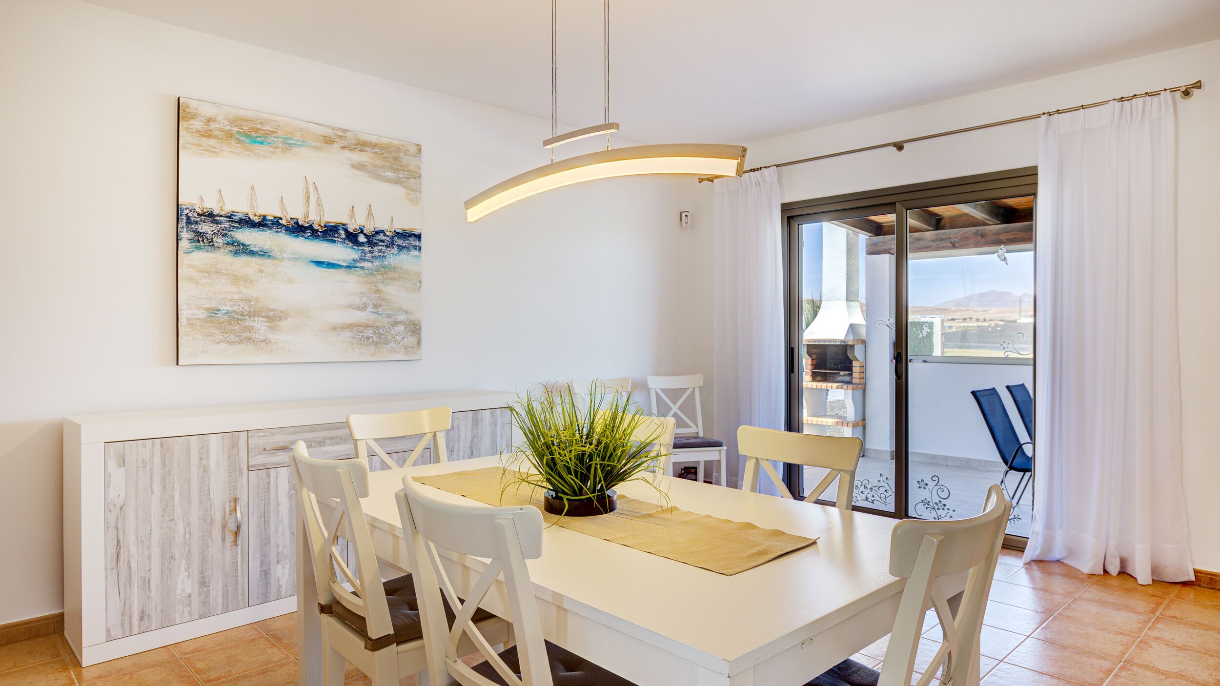 A dining area with a white table and chairs, a potted plant, and a painting of sailboats on the wall.
