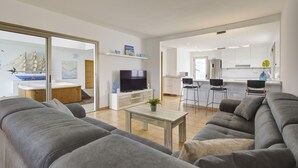 Een moderne woonkamer met een grijze bank, een televisie op een houten dressoir, en een keuken op de achtergrond.
