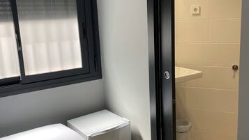 A small, modern bedroom with a bed, a white bedside table, a window, and a door leading to a bathroom.