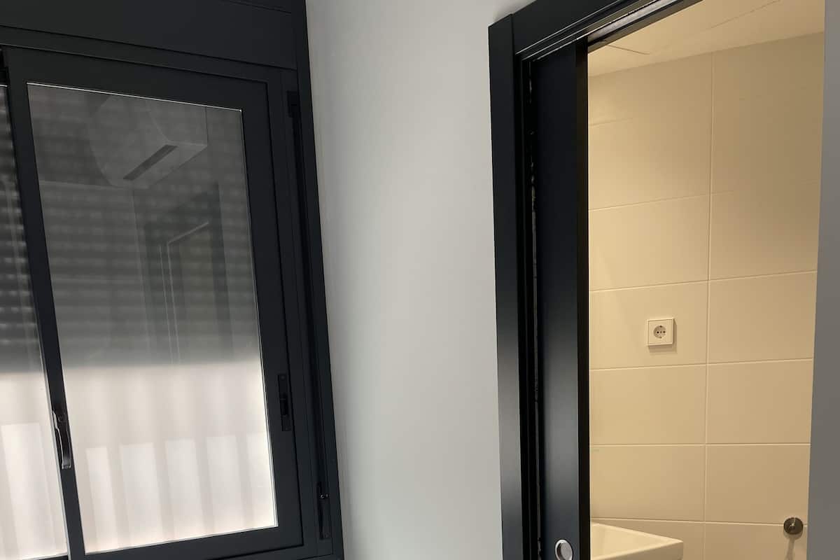 A small, modern room with a bed, a white bedside table, a window with blinds, and a door leading to a bathroom.