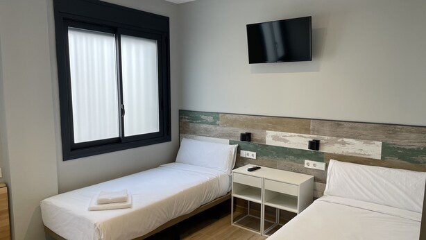 Une chambre d’hôtel avec un lit simple, une table de chevet, un téléviseur fixé au mur et une fenêtre avec des stores.