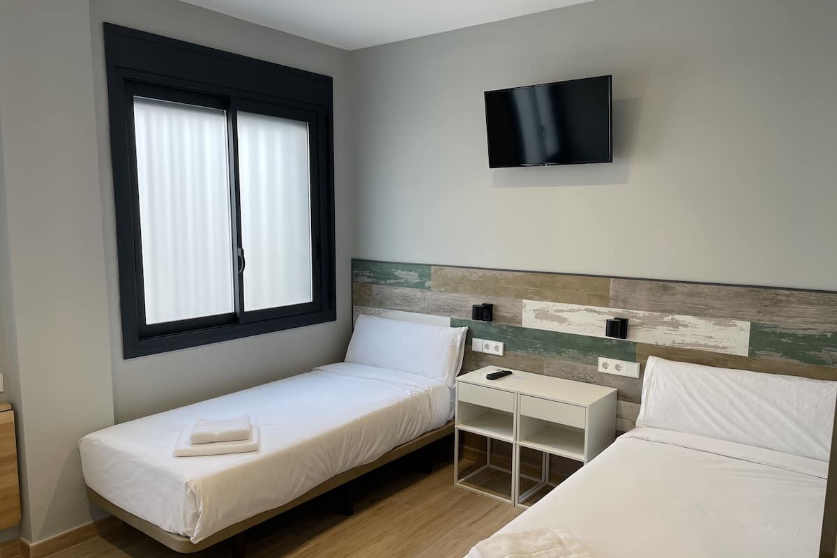 A hotel room with a single bed, a smaller bed, a TV mounted on the wall, a bedside table, and a window with blinds.