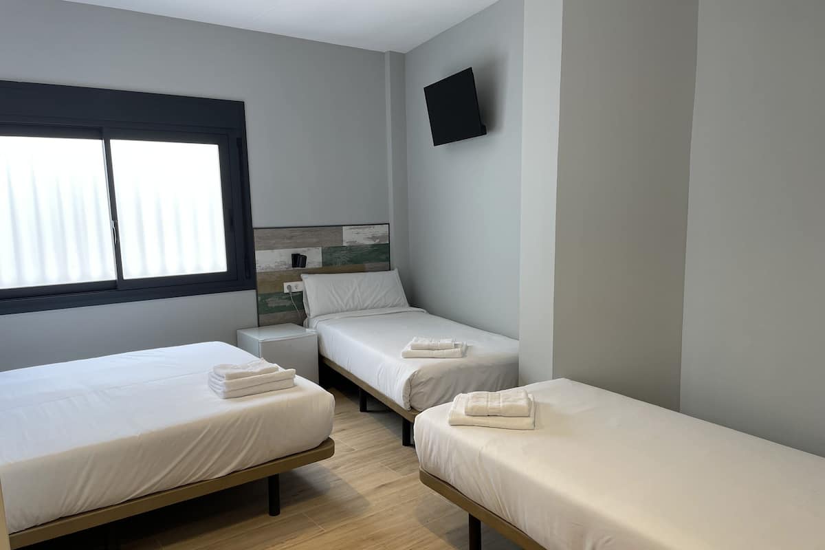 A room with three single beds, a small bedside table with a lamp, a window with blinds, and a flat-screen TV mounted on the wall.