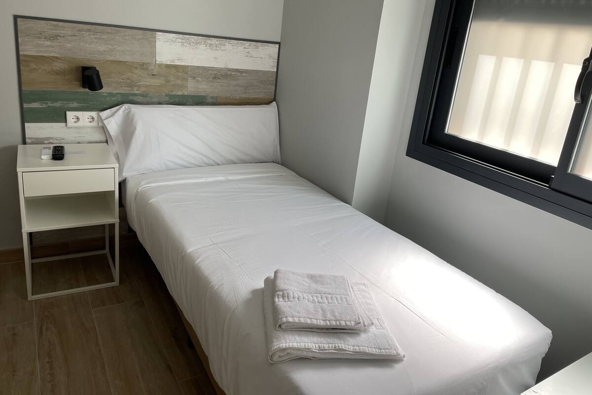A small, neatly made bed with white linens and a folded towel, a white nightstand with a remote control, and a wall-mounted power outlet.