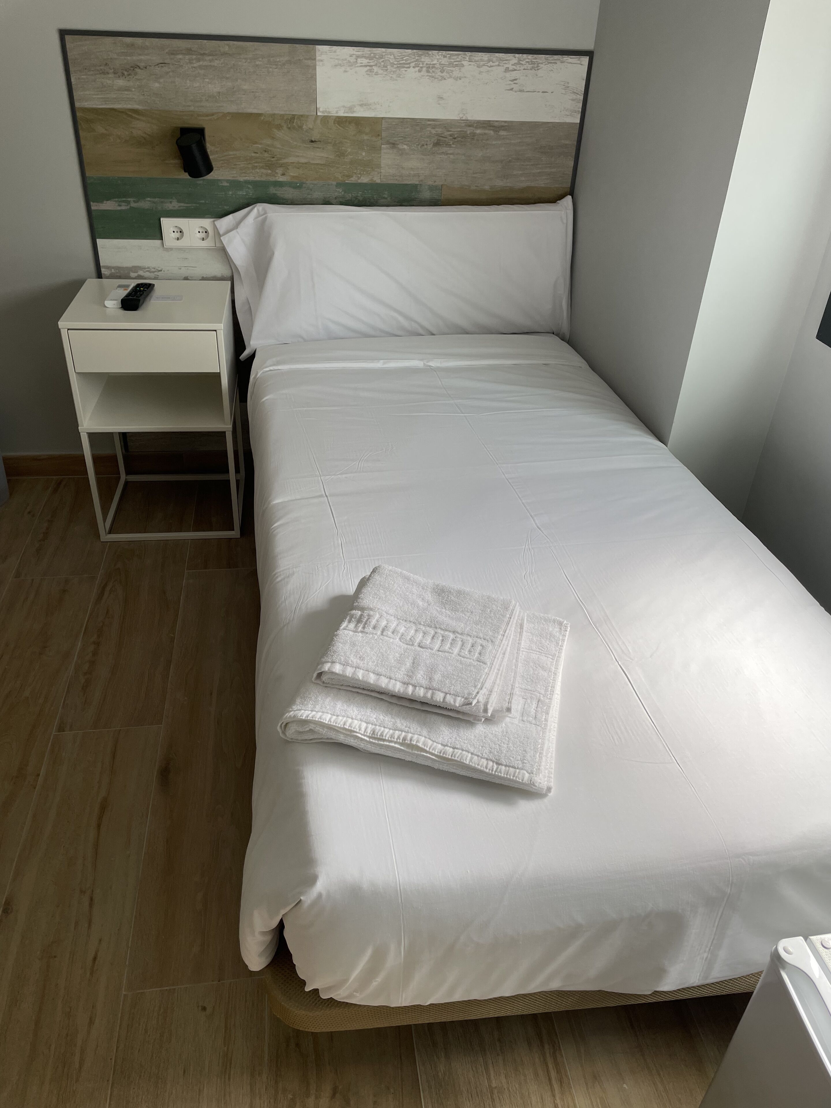 Un lit bien fait, avec du linge de lit blanc et deux serviettes pliées, une table de chevet avec une télécommande et un téléphone fixé au mur.