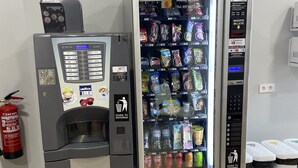 Un distributeur automatique proposant une variété de collations et de boissons, un terminal de paiement et une poubelle.