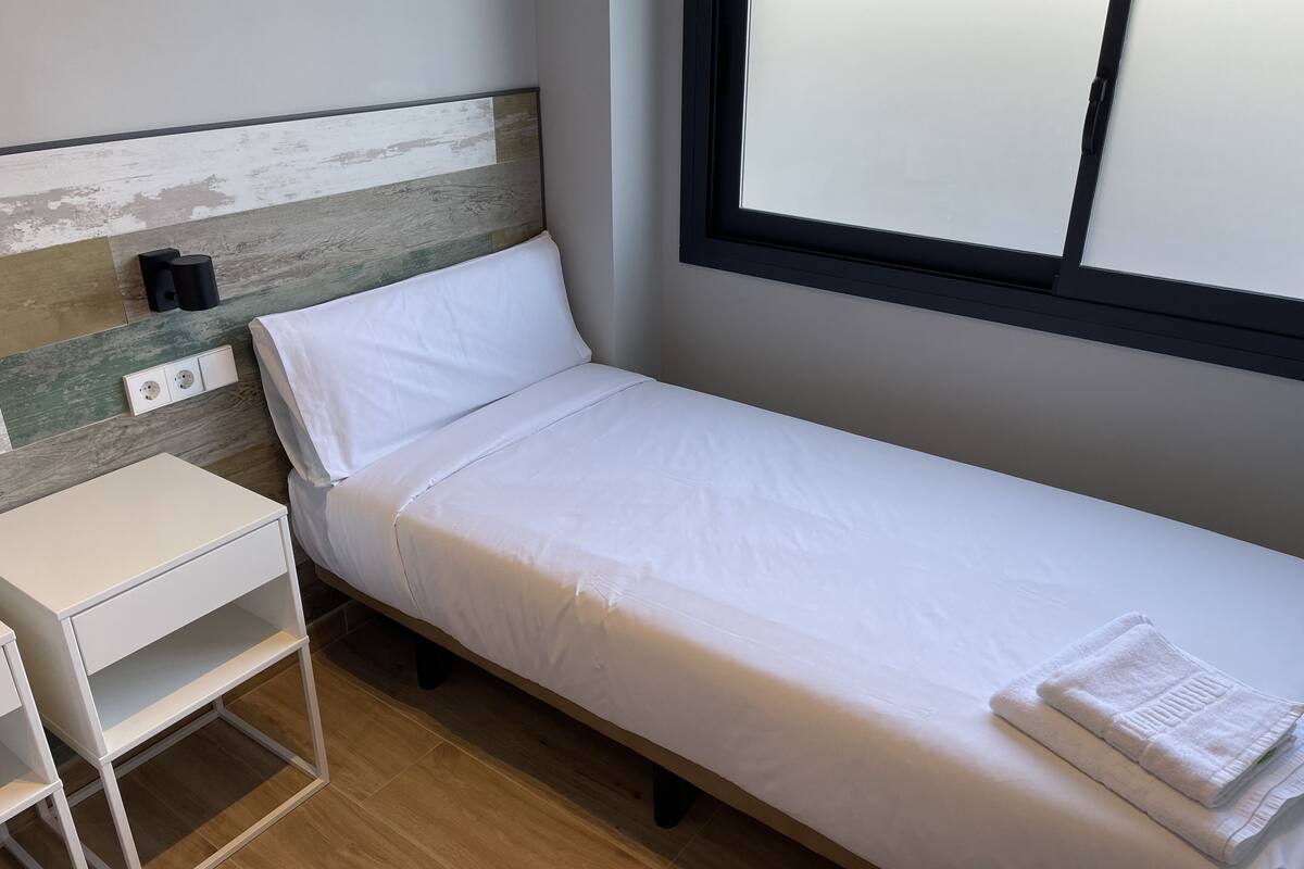 A single bed with white bedding, a small white desk with drawers, and a power outlet on the wall.