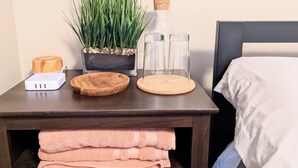 Une chambre d’hôtel bien rangée, avec un lit, une table de chevet, une plante en pot, une bouteille, des verres, un chargeur et des serviettes pliées.