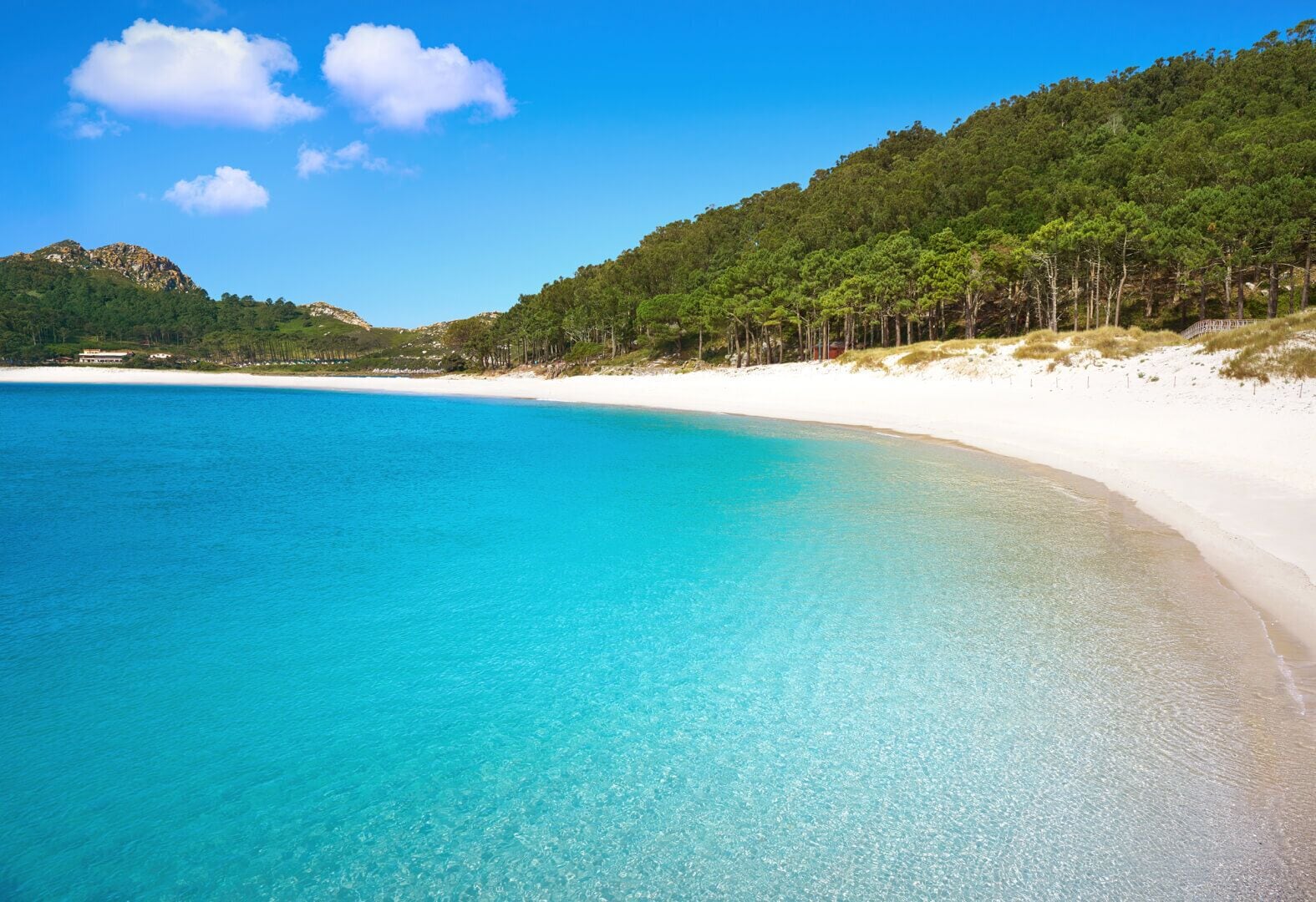A beach with clear turquoise water, white sand, and a forested hillside.