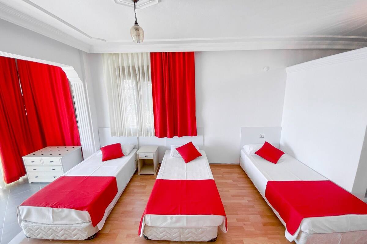 A hotel room with three single beds, each with a red blanket, white headboards, and a small bedside table. The room has white walls, a wooden floor, and a window with red curtains.