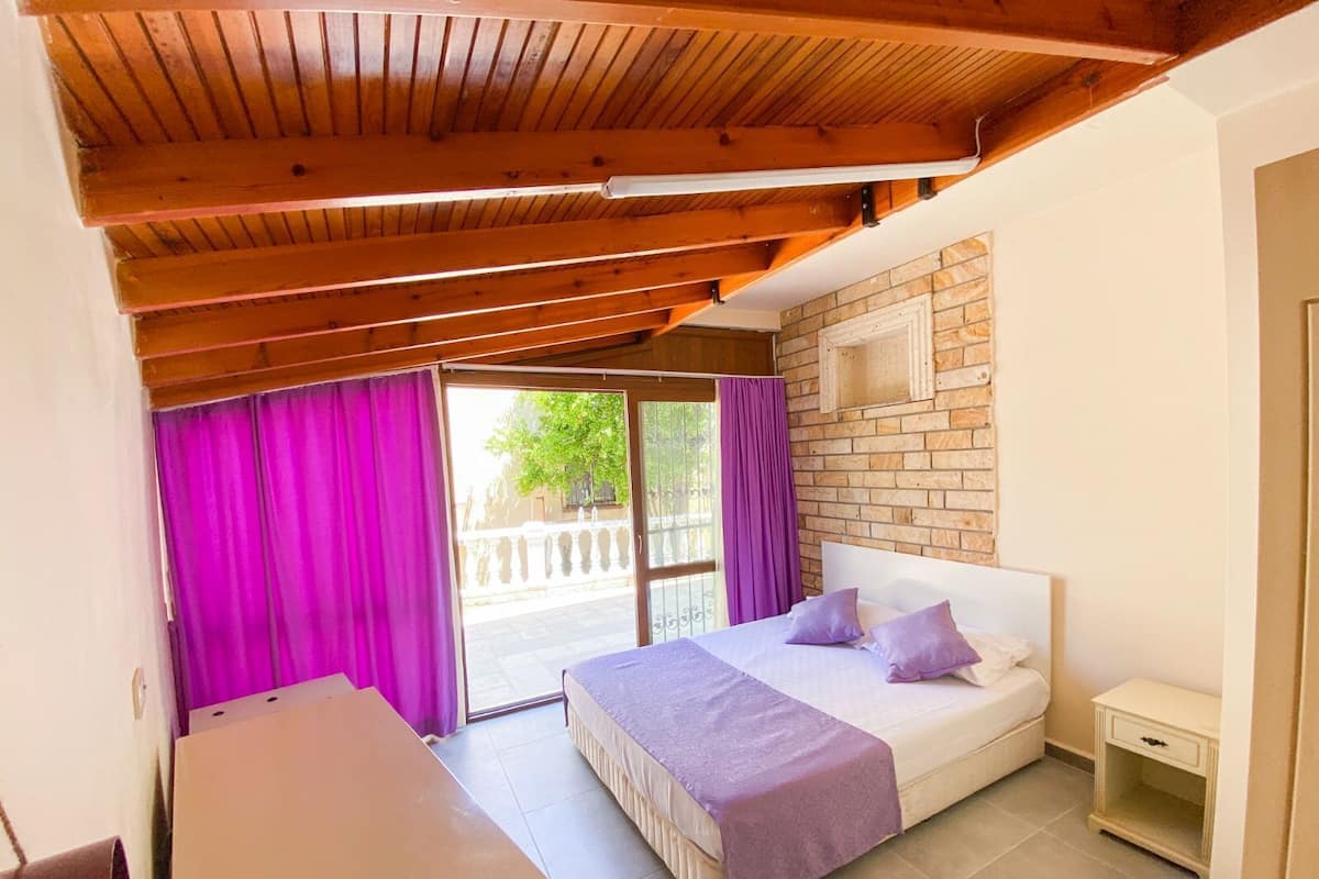 A bedroom with a wooden ceiling, a brick wall, a bed with a purple bedspread, and a small bedside table.