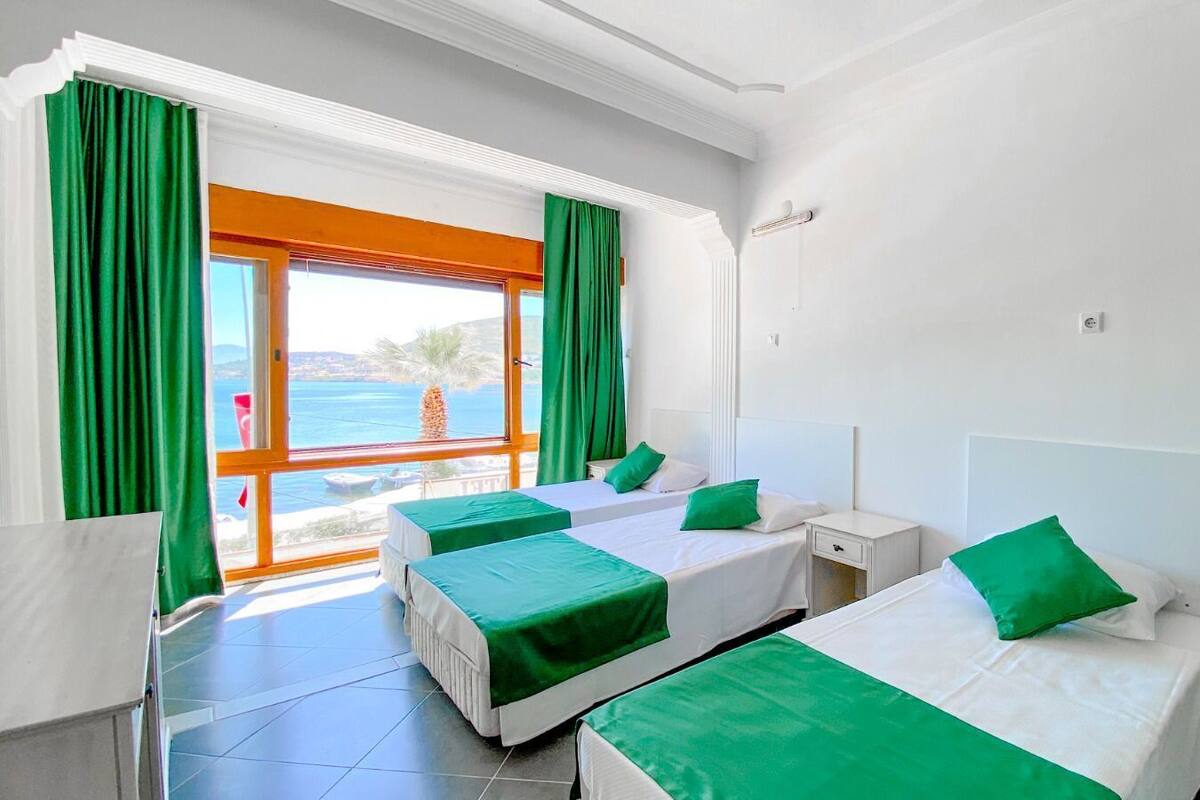 A hotel room with two beds, green bedspreads, a window with a view of the sea, and a door leading outside.