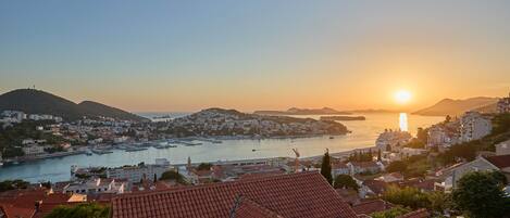 Eine Küstenstadtlandschaft mit Sonnenuntergang über dem Wasser, mit Wohnhäusern mit roten Ziegeldächern und einem Yachthafen.