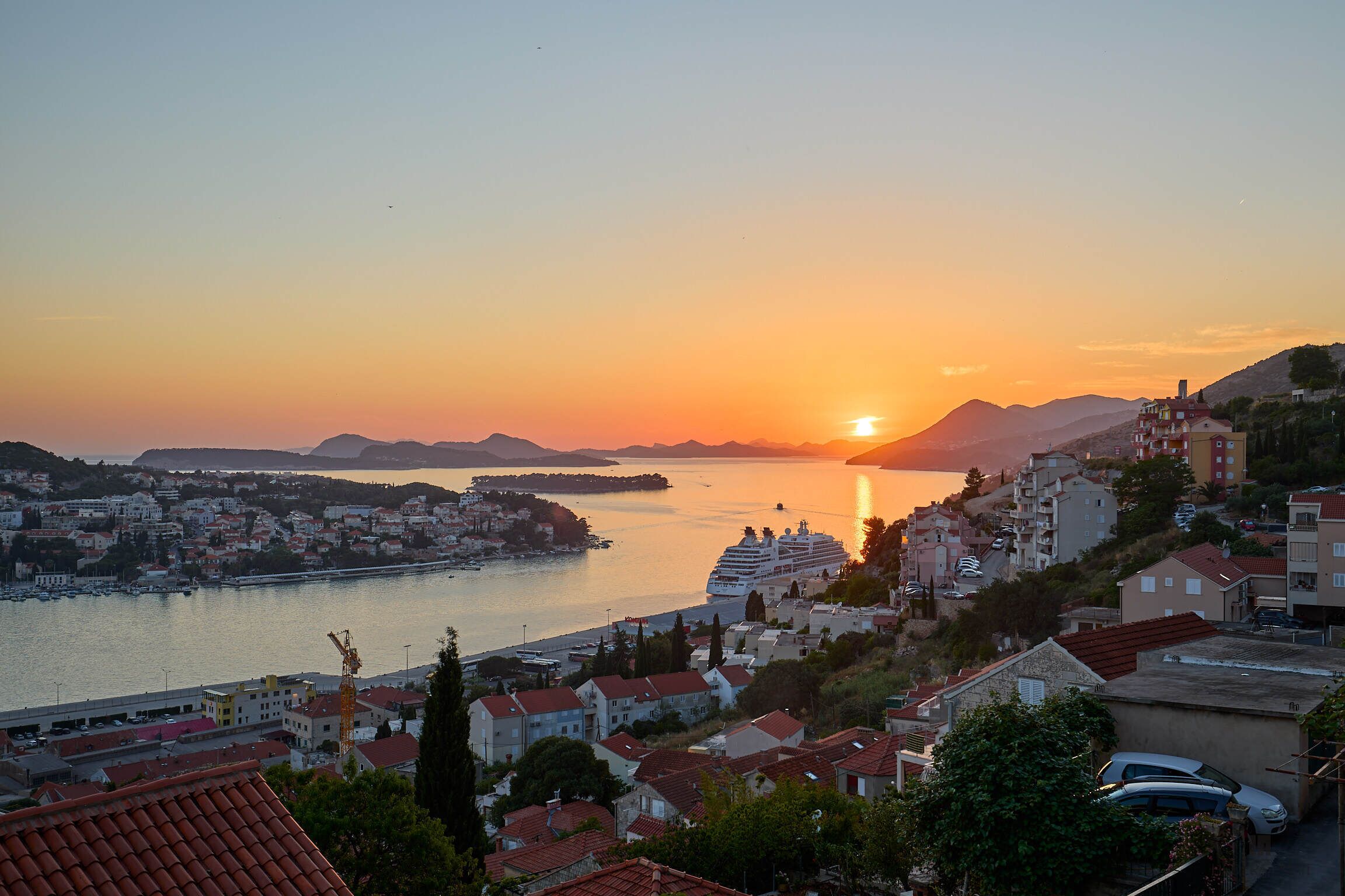 Um horizonte costeiro ao pôr do sol, com um cruzeiro atracado perto da praia, cercado por edifícios residenciais e colinas.